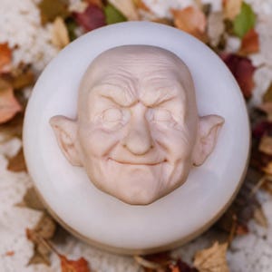 Peut inclure: Une sculpture de couleur chair pâle représentant un visage chauve et âgé avec des oreilles pointues et un sourire subtil, centrée sur une base sphérique blanche et lisse. Le visage présente des yeux fermés, un nez proéminent et des rides.