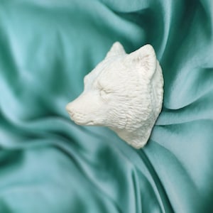 Puede incluir: Escultura de cerámica blanca de una cabeza de lobo con un diseño de pelaje texturizado. La escultura descansa sobre una tela de satén azul turquesa.