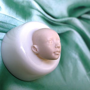 Pu&ograve; includere: Uno stampo in ceramica bianca con un viso di bambino scolpito in dettaglio. Lo stampo &egrave; su una superficie di tessuto verde.
