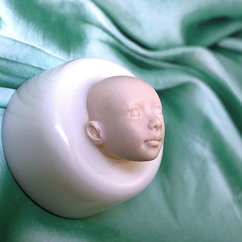 Pu&ograve; includere: Uno stampo in ceramica bianca con un viso di bambino scolpito in dettaglio. Lo stampo &egrave; su una superficie di tessuto verde.