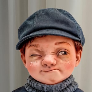 May include: A doll with red hair, wearing a blue denim newsboy cap and a blue turtleneck sweater. The doll has a mischievous expression, winking one eye and with a smudge on its nose.
