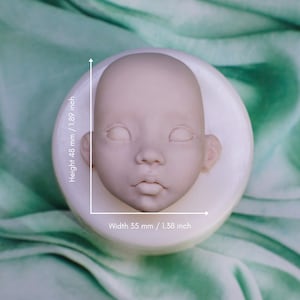 May include: A light beige doll head, 1.89 inches tall and 1.38 inches wide, with closed eyes, a small nose, and a slightly parted mouth. The head is set on a white, circular base, against a soft green background. This item is ideal for doll making.