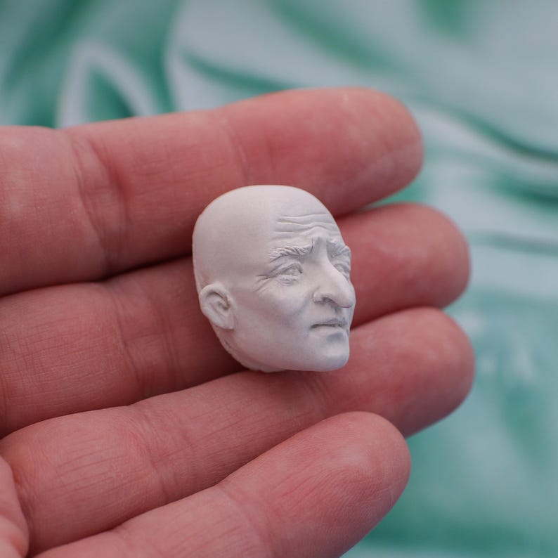 May include: A small, white, sculpted head of an elderly person with detailed facial features, including wrinkles and a prominent nose. The head is bald and appears to be made of a plaster or ceramic material. The sculpture is held in a person's hand.