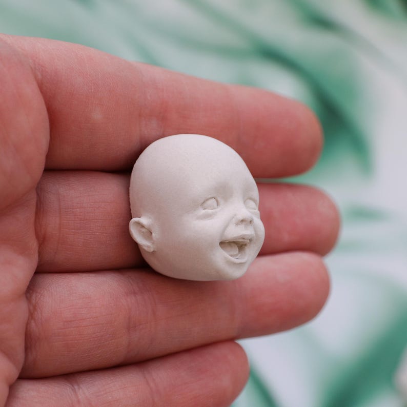 May include: A small, unpainted, white ceramic baby head with a smiling expression. The head features detailed facial features, including open eyes, a smiling mouth, and visible ears. The sculpture is held in a hand.