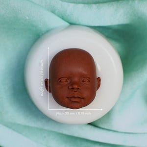 May include: A silicone mold for making a baby doll head. The mold is white and has a brown baby doll head inside. The mold is 2.5 inches tall and 0.78 inches wide.