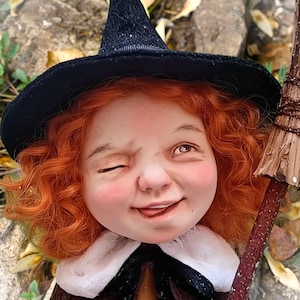 Könnte beinhalten: Eine skurrile Hexenpuppe mit roten Locken, rosigen Wangen und einem schwarzen Spitzhut. Sie zwinkert und streckt die Zunge heraus, während sie einen kleinen Besen hält. Die Puppe trägt einen weißen Kragen und ein braun-schwarzes Outfit.