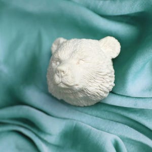 Op de afbeelding: Witte keramische berenkop sculptuur met een gesloten oog en een zachte, harige textuur. Het beeldje rust op een lichtblauwe stoffen achtergrond.