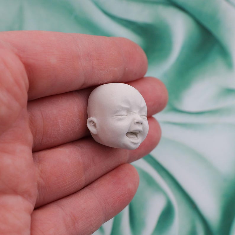 Peut inclure: Une petite t&ecirc;te de b&eacute;b&eacute; sculpt&eacute;e en blanc, les yeux ferm&eacute;s et la bouche ouverte, repr&eacute;sentant une expression de pleurs. La sculpture miniature est tenue dans une main, avec un arri&egrave;re-plan flou.