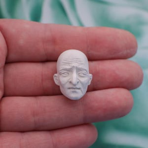 May include: A small, white, sculpted head of an elderly person with detailed facial features, including wrinkles and a bald head. The sculpture is held in a hand against a soft, green background.