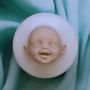 Könnte beinhalten: Eine kreisförmige, cremeweiße Silikonform mit einem detaillierten, dreidimensionalen Abdruck eines lächelnden Babygesichts. Die Form hat eine glatte, matte Oberflächenbeschaffenheit und dient zur Erstellung eines Reliefs eines Kinderkopfes mit offenen Augen.
