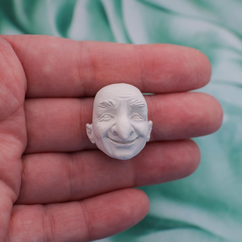 May include: A small, white, sculpted face with a smiling expression. The face has detailed features including a prominent nose, thin eyebrows, and a bald head. The sculpture is held in a person's hand.