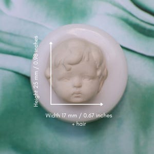 Può includere: Uno stampo in silicone bianco circolare raffigurante il volto scolpito di un bambino con capelli dettagliati. Lo stampo ha un'altezza di 25 mm e una larghezza di 17 mm più i capelli, come indicato dalle linee di misurazione bianche.