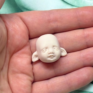 Puede incluir: Una pequeña cabeza de muñeca blanquecina con rasgos delicados, incluyendo ojos cerrados, una boca pequeña y orejas puntiagudas. La cabeza de la muñeca se sostiene en la mano de una persona, mostrando su tamaño y detalle. La cabeza parece estar hecha de un material liso.