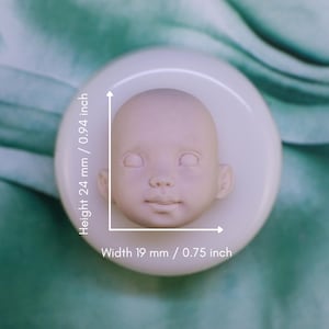 May include: A light beige doll head with a serene expression, measuring 0.94 inches in height and 0.75 inches in width. The doll head is set on a white, circular base. The image is set against a soft, green background.