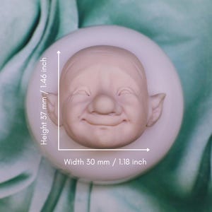 Könnte beinhalten: Eine hellbeige lächelnde Gesichtsskulptur mit spitzen Ohren, auf einem weißen kreisförmigen Sockel. Das Gesicht misst 37 mm in der Höhe und 30 mm in der Breite. Die Skulptur hat einen fröhlichen Ausdruck.