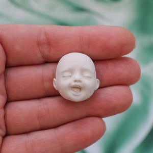 May include: A small, unpainted doll head with closed eyes and an open mouth, held in a person's hand. The doll head is a light, neutral color, and the facial features are delicately sculpted.
