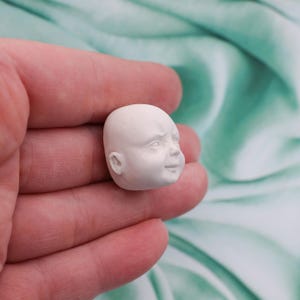 Peut inclure: Une petite t&ecirc;te de poup&eacute;e de b&eacute;b&eacute; blanche et non peinte. La t&ecirc;te est lisse et pr&eacute;sente des traits du visage d&eacute;taill&eacute;s, notamment un nez, une bouche et des oreilles. La t&ecirc;te de poup&eacute;e est tenue dans une main sur un fond doux de couleur sarcelle.