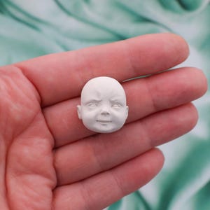 Peut inclure: Une petite t&ecirc;te de poup&eacute;e de b&eacute;b&eacute; sculpt&eacute;e en blanc, avec un l&eacute;ger froncement de sourcils et un l&eacute;ger sourire. La t&ecirc;te est lisse et d&eacute;taill&eacute;e, mettant en valeur les traits d'un jeune enfant. La t&ecirc;te de poup&eacute;e est une fourniture artisanale.