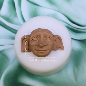 May include: A white silicone mold with a brown, detailed sculpture of a goblin face with large ears. The mold is 2.7 inches tall and 0.91 inches wide, excluding the ears.