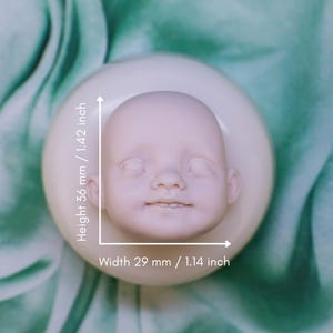 Könnte beinhalten: Ein hellrosa Baby-Puppenkopf mit geschlossenen Augen und einem leichten Lächeln. Der Kopf ist 36 mm hoch und 29 mm breit, auf einer weißen kreisförmigen Basis. Der Hintergrund ist ein weicher grüner Stoff.
