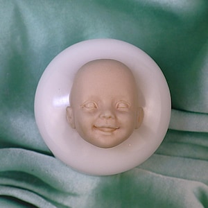 May include: A light beige baby face sculpture with a smiling expression, set within a white circular mold. The sculpture is centered on a green fabric background.