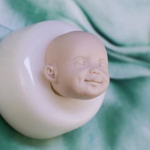 Może przedstawiać: Niemalowana głowa lalki dla dziecka z uśmiechniętą miną, umieszczona w białym, zaokrąglonym uchwycie. Głowa lalki ma szczegółowe cechy i jest umieszczona na tle turkusowej tkaniny.