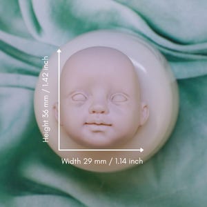 Puede incluir: Una cabeza de muñeca bebé sin pintar, de 36 mm de alto y 29 mm de ancho, colocada en una base redonda blanquecina. La cabeza presenta ojos cerrados, una nariz pequeña y una boca ligeramente entreabierta. El diseño general es suave y detallado.