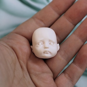 K&ouml;nnte beinhalten: Ein kleiner, blasser Puppenkopf mit matter Oberfl&auml;che und modellierten Gesichtsz&uuml;gen, darunter geschlossene Augen, eine kleine Nase und ein neutraler Mundausdruck. Der Kopf ist glatt, abgerundet und haarlos, was ein realistisches Aussehen verleiht.
