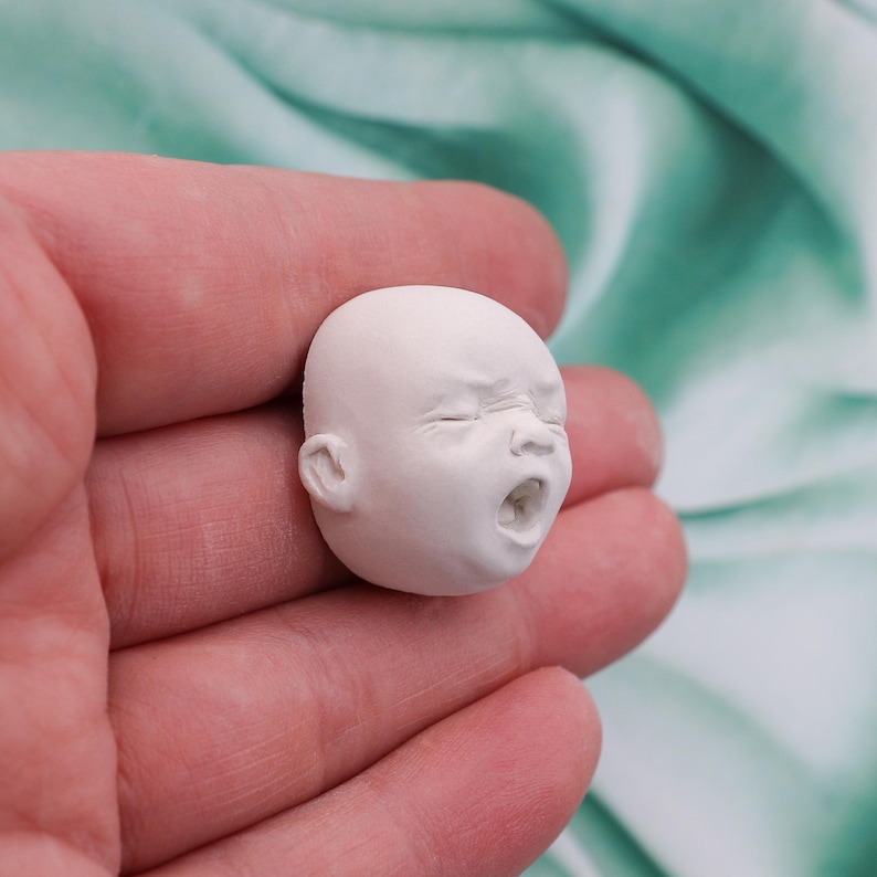 Peut inclure: Une petite t&ecirc;te de b&eacute;b&eacute; sculpt&eacute;e en blanc, avec une expression de d&eacute;tresse. La bouche est ouverte en pleurs et les yeux sont pliss&eacute;s. La sculpture miniature est tenue dans une main sur un fond vert clair et doux.
