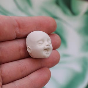May include: A small, unpainted, white baby doll head with closed eyes and an open mouth, sculpted to appear as if laughing. The head is smooth and detailed, showcasing the features of a smiling infant. Ideal for crafting or doll making.