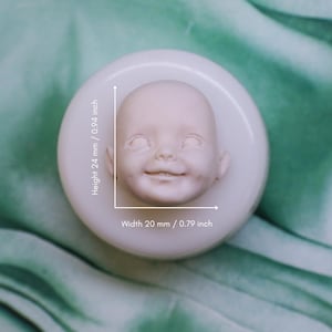 May include: A light-colored, smiling baby face mold, 0.94 inches tall and 0.79 inches wide, sits on a white circular base. The baby face has detailed features and a cheerful expression. This mold is likely for crafting or doll making.