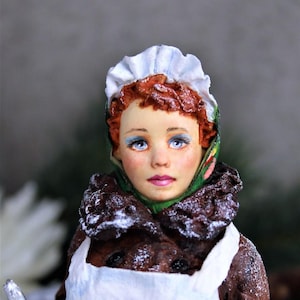 May include: A porcelain doll with red hair, wearing a white bonnet and a green and red scarf. The doll is dressed in a white apron and a brown coat with white powder on it.