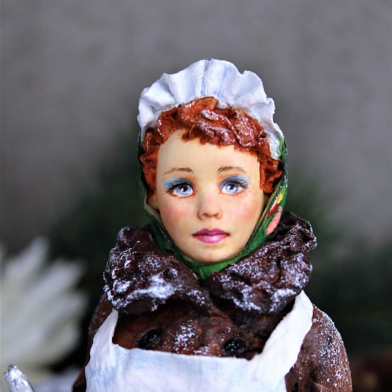 May include: A porcelain doll with red hair, wearing a white bonnet and a green and red scarf. The doll is dressed in a white apron and a brown coat with white powder on it.