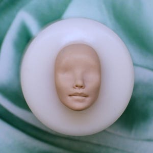 Può includere: Uno stampo in silicone bianco con un viso dettagliato scolpito al suo interno. Il viso è realizzato in un materiale marrone chiaro e ha un'espressione neutra.
