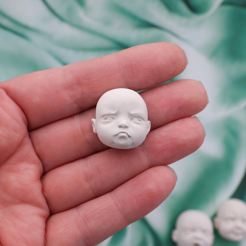 Peut inclure: Une petite t&ecirc;te de poup&eacute;e blanche, non peinte, avec une expression neutre. La t&ecirc;te de poup&eacute;e a les yeux ferm&eacute;s, un petit nez et une bouche l&eacute;g&egrave;rement tombante. La t&ecirc;te est tenue dans une main sur un fond vert et blanc doux.