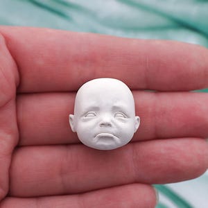 Peut inclure: Une petite t&ecirc;te de poup&eacute;e de b&eacute;b&eacute; sculpt&eacute;e en blanc, avec une expression triste. La t&ecirc;te a les yeux ferm&eacute;s, une bouche tombante et des traits d&eacute;taill&eacute;s. La t&ecirc;te de poup&eacute;e est tenue dans la main d'une personne.