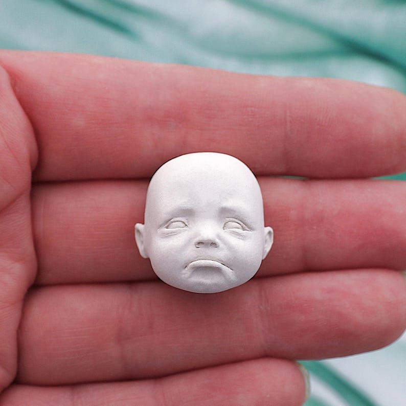 Peut inclure: Une petite t&ecirc;te de poup&eacute;e de b&eacute;b&eacute; sculpt&eacute;e en blanc, avec une expression triste. La t&ecirc;te a les yeux ferm&eacute;s, une bouche tombante et des traits d&eacute;taill&eacute;s. La t&ecirc;te de poup&eacute;e est tenue dans la main d'une personne.