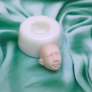 Pu&ograve; includere: Uno stampo in silicone bianco per realizzare una testa di bambola, con una testa di bambola realizzata in materiale rosa chiaro accanto.