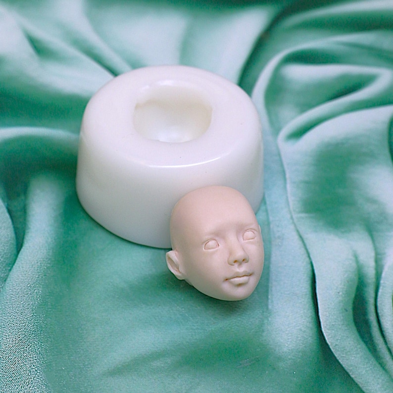Pu&ograve; includere: Uno stampo in silicone bianco per realizzare una testa di bambola, con una testa di bambola realizzata in materiale rosa chiaro accanto.