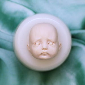 K&ouml;nnte beinhalten: Eine cremewei&szlig;e Silikonform mit dem detailliert geformten Gesicht eines Babys mit geschlossenen Augen, einer kleinen Nase und einem neutralen Ausdruck. Die Form ist kreisf&ouml;rmig und hat eine glatte, matte Oberfl&auml;che, bestimmt f&uuml;r Puppenk&ouml;pfe.