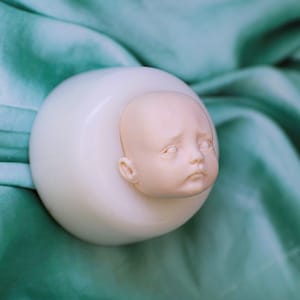 K&ouml;nnte beinhalten: Ein glattes, cremewei&szlig;es, kugelf&ouml;rmiges Objekt mit einem realistischen, blassfarbenen Puppenkopf, der aus der Mitte herausragt. Der Puppenkopf hat geschlossene Augen, eine kleine Nase und einen neutralen Ausdruck, nahtlos in die runde Form.