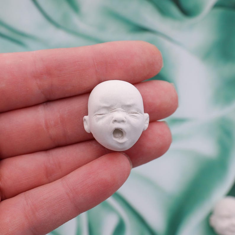 Peut inclure: Une petite t&ecirc;te de b&eacute;b&eacute; sculpt&eacute;e en blanc, avec les yeux ferm&eacute;s et la bouche ouverte, tenue dans une main. La t&ecirc;te est d&eacute;taill&eacute;e avec des rides et des traits du visage, sugg&eacute;rant une expression de d&eacute;tresse. La sculpture est probablement destin&eacute;e &agrave; l'artisanat ou &agrave; la fabrication de poup&eacute;es.