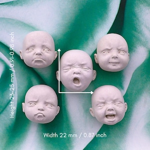 Puede incluir: Cinco pequeñas cabezas de muñeca sin pintar con diversas expresiones faciales, incluyendo llanto, bostezo y sonrisa. Las cabezas miden aproximadamente 22 mm de ancho y 23-25 mm de alto. El fondo es un patrón abstracto verde y blanco.
