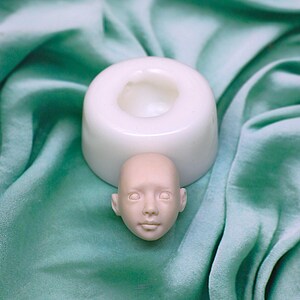 Pu&ograve; includere: Uno stampo in silicone bianco per realizzare una testa di bambola. Lo stampo &egrave; appoggiato su una superficie di tessuto verde. Una piccola testa di bambola non dipinta &egrave; appoggiata accanto allo stampo.
