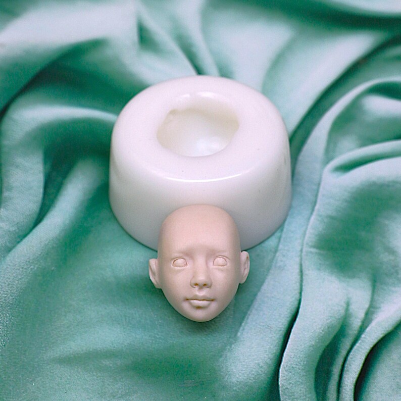 Pu&ograve; includere: Uno stampo in silicone bianco per realizzare una testa di bambola. Lo stampo &egrave; appoggiato su una superficie di tessuto verde. Una piccola testa di bambola non dipinta &egrave; appoggiata accanto allo stampo.