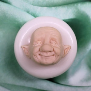May include: A white silicone mold of a smiling, elderly man's face with large ears. The mold is on a white surface.