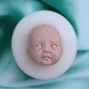 Puede incluir: Un molde de silicona para crear una cabeza de muñeca bebé. El molde es blanco y tiene una impresión detallada de la cara de un bebé con los ojos cerrados.