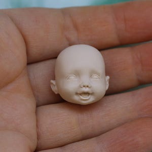 May include: A close-up of a small, unpainted, porcelain doll head with a smiling expression. The head is sculpted with realistic features, including eyes, a nose, and a mouth.