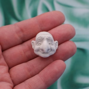 May include: A small, off-white sculpture of a smiling face with a prominent nose, furrowed brow, and small ears. The face has a whimsical, aged appearance, suggesting a character from a fantasy or folklore theme. The sculpture is held in a hand.
