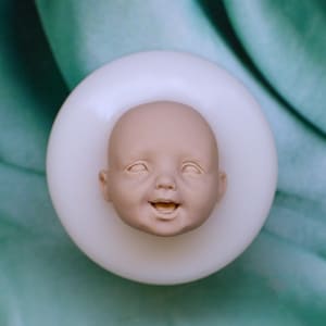 Könnte beinhalten: Eine Silikonform mit einem detaillierten, dreidimensionalen Babygesicht mit lächelndem Ausdruck, offenem Mund und geschlossenen Augen. Die Form ist kreisförmig, weiß und undurchsichtig und für Gussarbeiten mit einer glatten, matten Oberfläche.